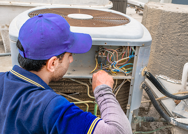 A man in a blue baseball cap operates on an air conditioning unit outdoors.