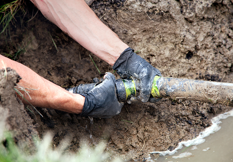 the arms of a technician fixing a broken sewer line in the mud