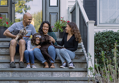 a hispanic family consisting of a dad, mom, two daughters and two dogs sitting on the font steps of their home